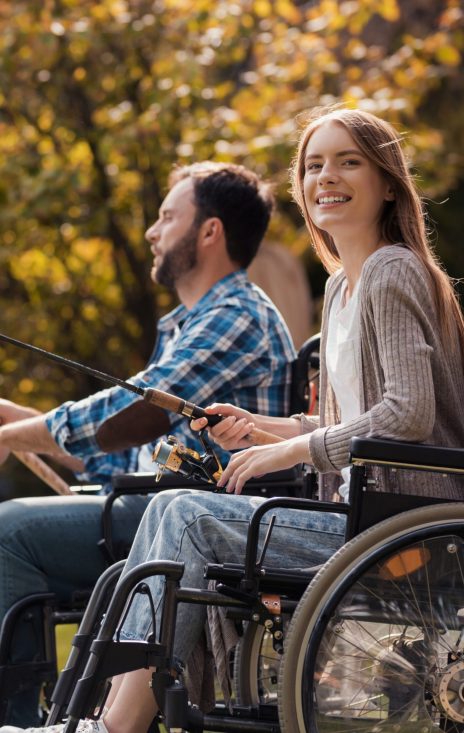 A woman and a man in wheelchairs are sitting on the lake shore. They have spinnings in their hands and they are fishing. They are smiling. Behind them are beautiful green trees and a park.