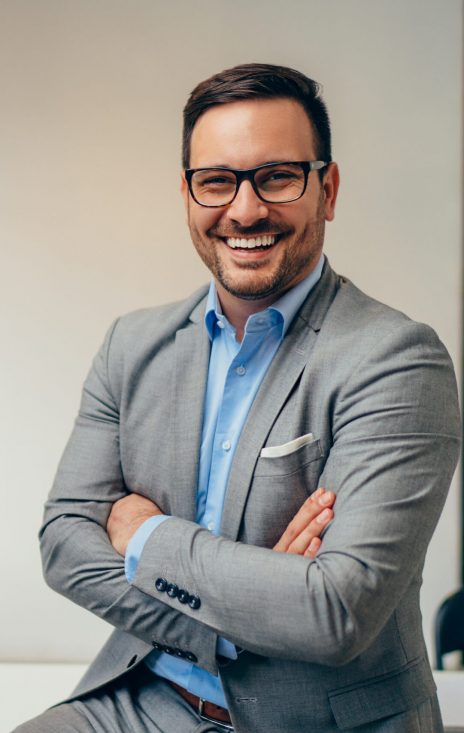 Portrait of young happy businessman wearing grey suit and blue shirt standing in his office and smiling with arms crossed